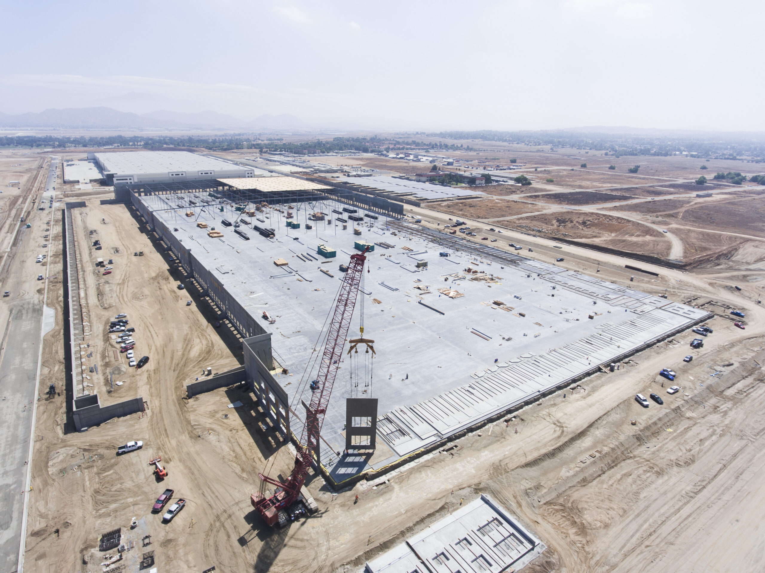 Aerial view of a construction site with a crane lifting a concrete wall panel beside a large building slab.